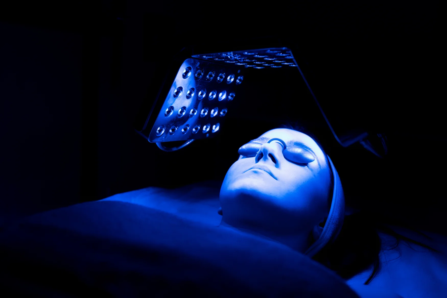 A woman wearing protective glasses is lying under a blue light machine.
